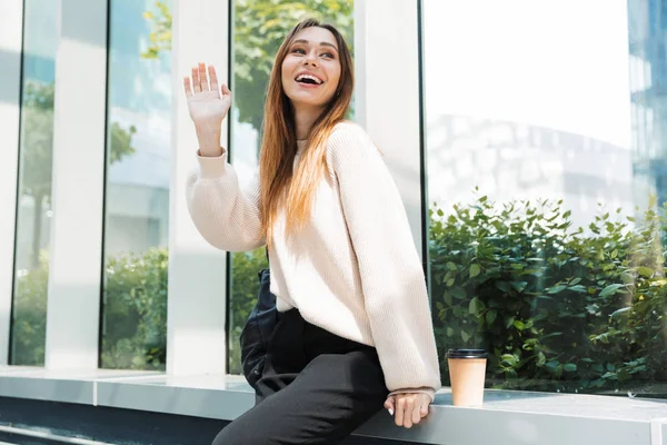 Photo of caucasian charming woman smiling and waving her hand while sitting near window in building indoors