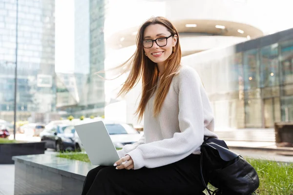 Belle jeune femme d'affaires heureuse posant assis à l'extérieur près du centre d'affaires portant des lunettes à l'aide d'un ordinateur portable . — Photo