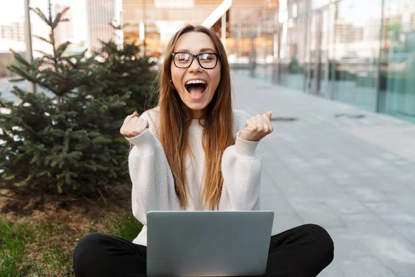 Hermosa mujer de negocios feliz emocionado impactado joven posando sentado al aire libre cerca de centro de negocios con gafas usando ordenador portátil hacer gesto ganador . — Foto de Stock