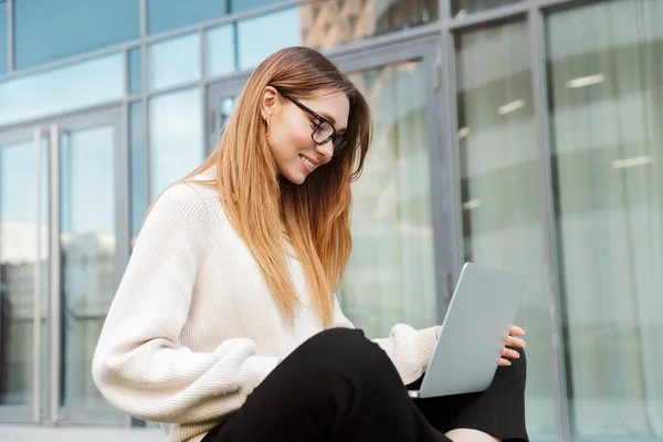 Güzel genç mutlu iş kadını dizüstü bilgisayar kullanarak gözlük takarak iş merkezi yakınında açık havada oturan poz. — Stok fotoğraf