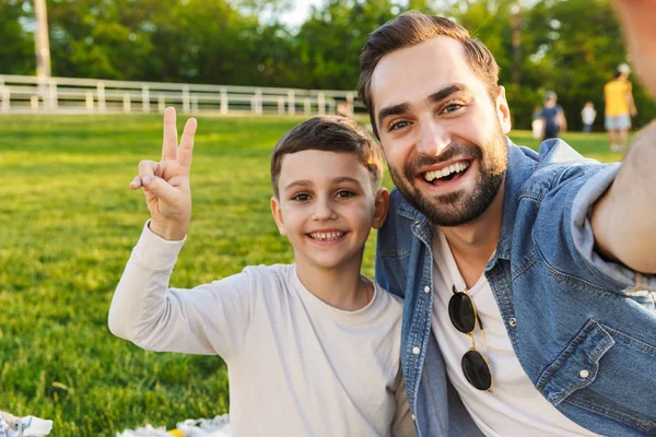 Fiatalember szórakozik az ő kis testvére vagy fia outoors a parkban gyönyörű zöld fű hogy a selfie a kamera. — Stock Fotó