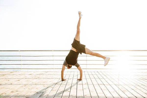 Sterke jonge sport man op het strand in de buurt van zee staande op armen handen maken yoga oefeningen. — Stockfoto