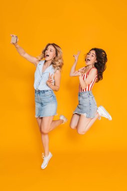 İki mutlu sarışın ve esmer kadın portresi cep telefonu selfie fotoğraf çekerken barış şarkı ve atlama gesturing