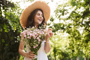 Yeşil parkta çiçekler tutan şapka yürüyüş Heyecanlı mutlu genç güzel kadın outoors.
