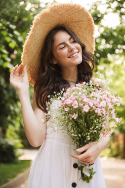 Yeşil parkta çiçekler tutan şapka yürüyüş Heyecanlı mutlu genç güzel kadın outoors.