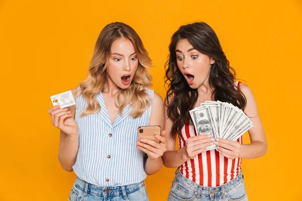 Porträt schockierter hübscher Frauen in Sommerkleidung mit Handy, während sie Kreditkarten- und Geldscheine in der Hand halten — Stockfoto