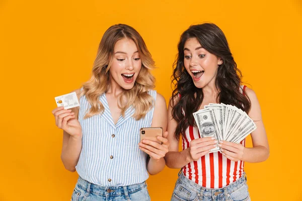 Porträt von entzückten hübschen Frauen in Sommerkleidung mit Handy, während sie Kreditkarten- und Geldscheine in der Hand halten — Stockfoto