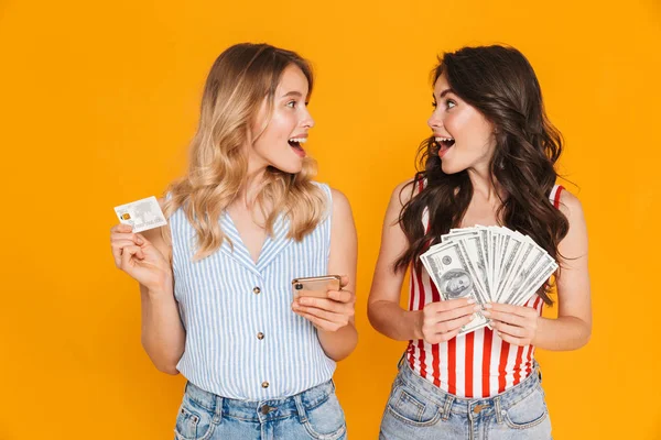 Porträt von entzückten süßen Frauen in Sommerkleidung mit Handy, während sie Kreditkarten- und Geldscheine in der Hand halten — Stockfoto