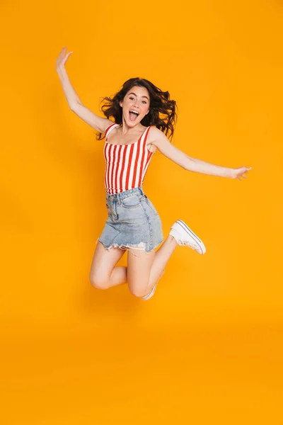 Retrato de mujer morena encantada en falda de mezclilla saltando con los brazos levantados —  Fotos de Stock