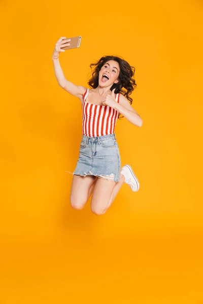 Retrato de mujer morena excitada en falda de mezclilla saltando y mostrando el pulgar hacia arriba mientras toma selfie en el teléfono celular —  Fotos de Stock