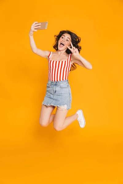 Retrato de mujer morena sorprendida en falda de mezclilla saltando y mostrando la paz cantando mientras toma selfie en el teléfono celular —  Fotos de Stock