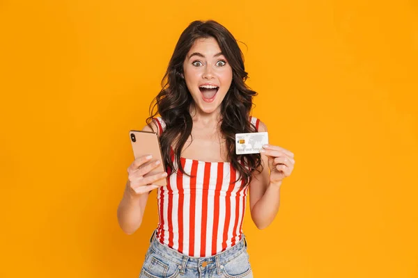 Jovem feliz menina emocional isolado sobre fundo de parede amarela usando telefone celular segurando cartão de crédito . — Fotografia de Stock