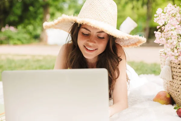 Photo of elegant young woman wearing straw hat using laptop while having picnic in green park — Stock Photo, Image