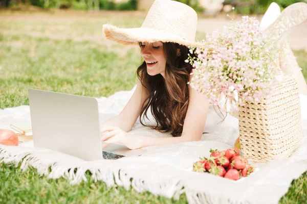 Photo of joyous young woman wearing straw hat typing on laptop while having picnic in green park — Stock Photo, Image