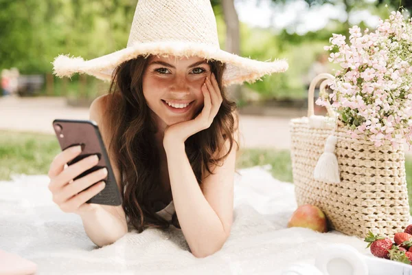 Φωτογραφία της καυκάσιας νεαρής γυναίκας φορώντας ψάθινο καπέλο κρατώντας smartphone ενώ έχει πικνίκ σε πράσινο πάρκο — Φωτογραφία Αρχείου