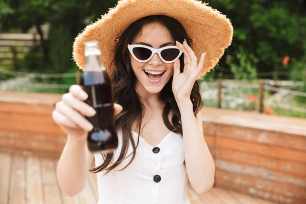 Opgewonden gelukkig jonge mooie vrouw outoors in Park poseren in hoed houden soda. — Stockfoto