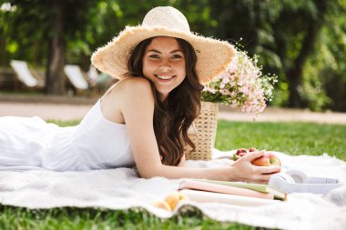 Piknik yaparken ve yeşil parkta battaniye üzerinde yatarken hasır şapka giyen sevimli genç kadının fotoğrafı