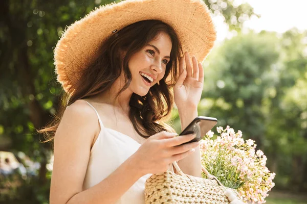 Emocionado feliz joven bonita mujer salidas en verde parque caminando en sombrero usando el teléfono móvil . — Foto de Stock