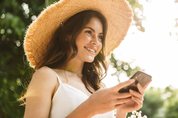 Foto de mujer caucásica elegante feliz usando sombrero de paja usando el teléfono celular mientras camina en el parque verde — Foto de Stock
