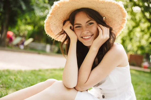 Foto einer fröhlichen jungen Frau mit Strohhut, die lächelt, während sie im grünen Park auf Gras sitzt — Stockfoto