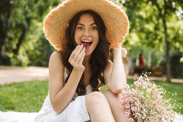 Aufgeregt glücklich junge hübsche Frau outoors in grünen Park in Hut sitzt auf Gras essen Erdbeere. — Stockfoto