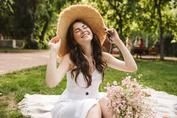 Emocionado feliz joven bonita mujer salidas en verde parque en sombrero sentado en la hierba usando el teléfono móvil escuchar música . —  Fotos de Stock
