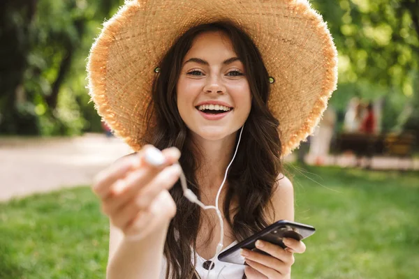 Aufgeregten fröhlichen jungen hübschen Frau outoors in grünen Park in Hut sitzt auf Gras mit Handy Musik hören. — Stockfoto