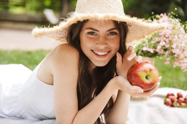 Photo of beautiful young woman wearing straw hat eating apple while having picnic in green park — Stock Photo, Image