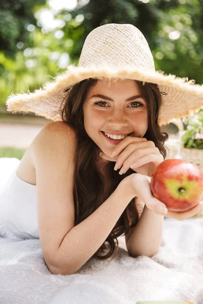 Ενθουσιασμένος ευτυχής νεαρή όμορφη γυναίκα σε πράσινο πάρκο στο καπέλο κάθεται στο γρασίδι τρώνε μήλο. — Φωτογραφία Αρχείου