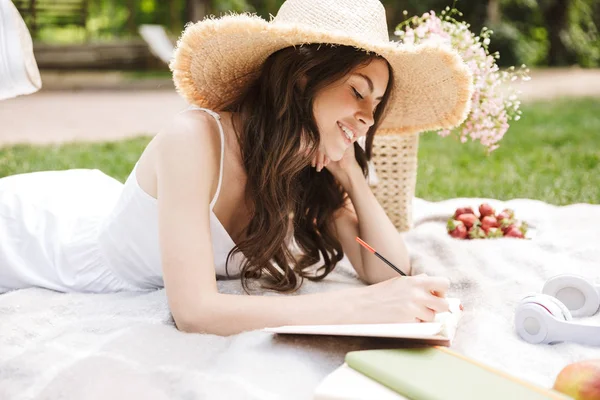 Foto di elegante giovane donna che indossa cappello di paglia annotando note al diario mentre fa un picnic nel parco verde — Foto Stock