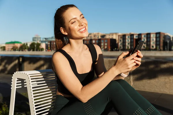 Image of joyful sporty woman holding smartphone and sitting on c — Stock Photo, Image