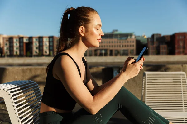 Imagem da mulher desportiva iluminada pelo sol segurando smartphone e sentado em c — Fotografia de Stock