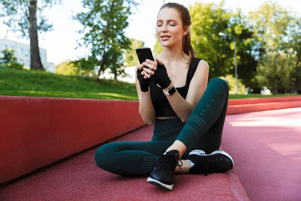 Fotografie atletické mladé ženy držící smartphone při sezení o — Stock fotografie