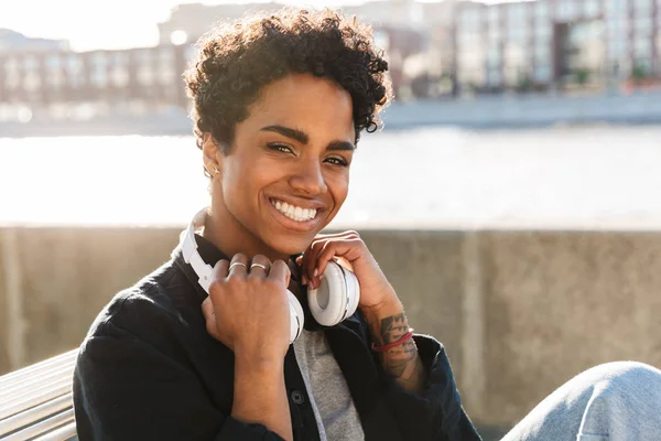 Porträtt närbild av glada African American Holding hörlurar när du sitter på bänken nära Riverfront — Stockfoto