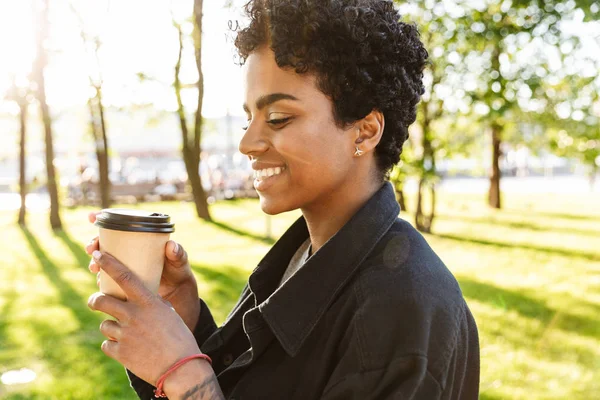 Fotografie atraktivní africké americké ženy, která drží a pije kávu z plastového kelímku při chůzi v městském parku — Stock fotografie