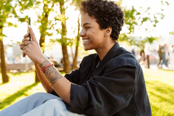 Fotografie spokojena s africkou americkou ženou, která si při sezení na lavičce v městském parku vydělala fotografii na telefonu — Stock fotografie