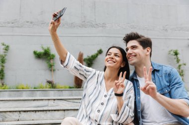 Gülümseyen neşeli genç sevgi dolu çift açık havada cep telefonu ile selfie çekmek.