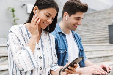 Mutlu genç şaşırtıcı sevgi dolu çift iş adamları iş arkadaşları cep telefonu ve dizüstü bilgisayar kulaklık ile müzik dinleme kullanarak adımlarla dışında açık havada.