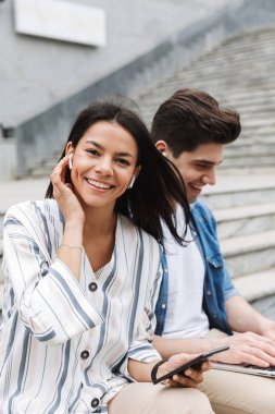 Mutlu genç şaşırtıcı sevgi dolu çift iş adamları iş arkadaşları cep telefonu ve dizüstü bilgisayar kulaklık ile müzik dinleme kullanarak adımlarla dışında açık havada.