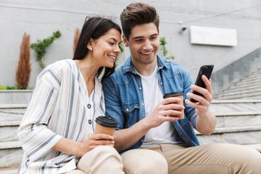 Neşeli çift paket kahve içme ve şehir merdivenlerinde açık havada akıllı telefon kullanarak Görüntü