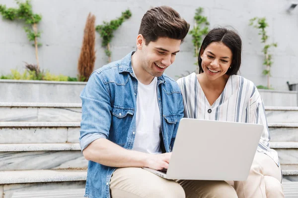 Imagen de una joven pareja sonriendo y usando un portátil mientras está sentada en el banco cerca de escaleras al aire libre — Foto de Stock