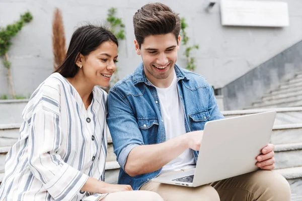 Imagen de pareja morena sonriendo y mirando al portátil mientras está sentada en el banco cerca de escaleras al aire libre — Foto de Stock