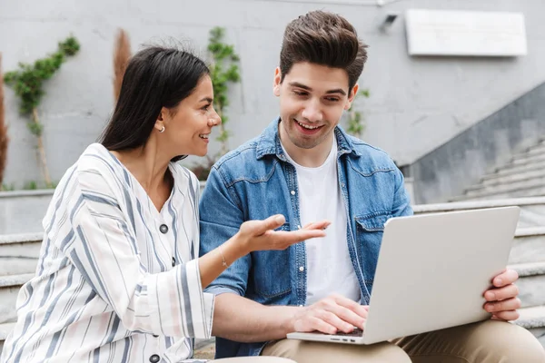 Beeld van brunette paar praten en het gebruik van laptop terwijl zittend op de Bank in de buurt van trappen buitenshuis — Stockfoto