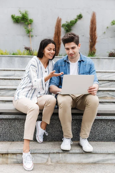 Imagen de pareja atractiva hablando y utilizando el ordenador portátil mientras está sentado en el banco cerca de escaleras al aire libre — Foto de Stock