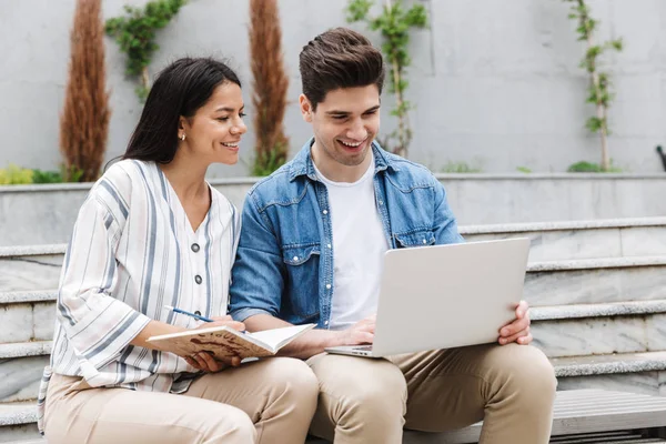 Imagen de una pareja sonriente usando un portátil y escribiendo en un diario mientras está sentada en un banco cerca de escaleras al aire libre — Foto de Stock