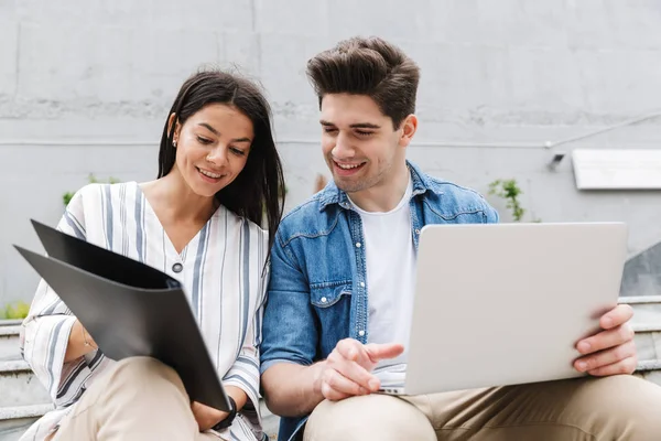 Счастливая молодая удивительная влюбленная пара бизнесменов использует ноутбук, держа в руках папку для чтения документов . — стоковое фото