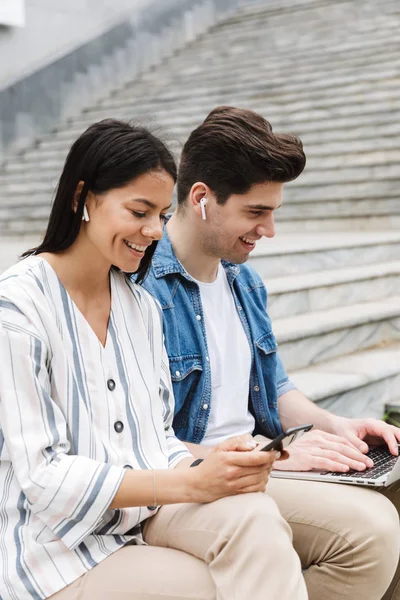 Mutlu genç şaşırtıcı sevgi dolu çift iş adamları iş arkadaşları cep telefonu ve dizüstü bilgisayar kulaklık ile müzik dinleme kullanarak adımlarla dışında açık havada.