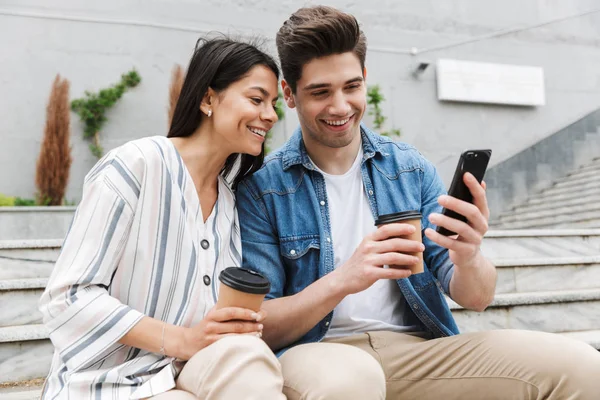 Afbeelding van vrolijke paar drinken afhalen koffie en het gebruik van smartphone op de stad trappen buitenshuis — Stockfoto