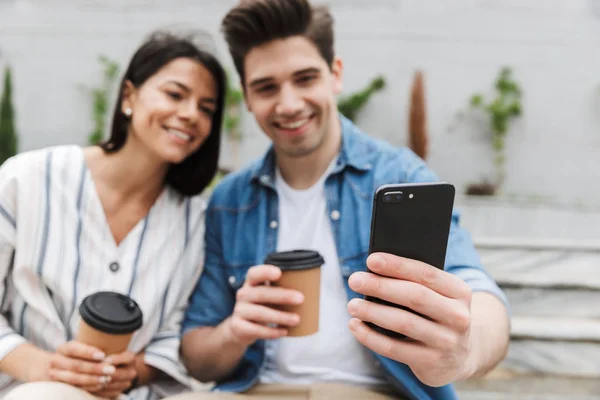 Verschwommenes Bild eines jungen Paares, das Kaffee zum Mitnehmen trinkt und sein Smartphone auf der Treppe der Stadt im Freien benutzt — Stockfoto