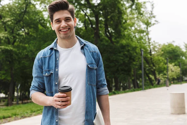 Image of handsome young man drinking takeaway coffee and holding laptop while walking through green park — Stock Photo, Image